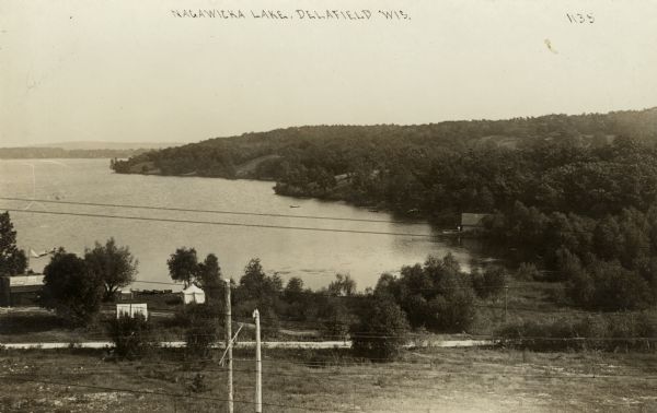 Elevated view from hill looking towards Nagawicka Lake. There is a road and buildings along the shoreline at the bottom of the hill. There are a number of boats on the lake. Power lines are in the foreground. Caption reads: "Nagawicka Lake, Delafield, Wis." 
