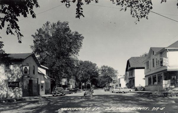 Black and white photographic postcard of Milwaukee Street, with storefronts, parked cars and a small monument in the middle of the street. Caption reads: "Milwaukee Street, Delafield, Wis."