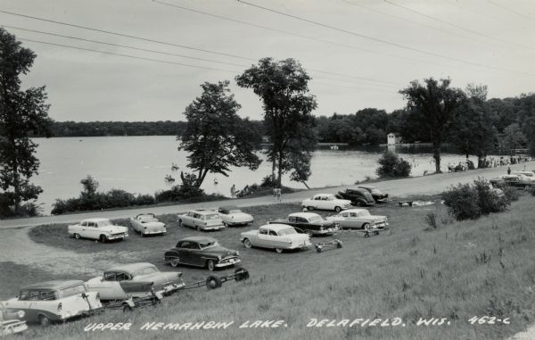 Black and white photographic postcard of Upper Nemahbin Lake, with parked cars at the base of a hill in the foreground, and people boating and swimming. Caption reads: "Upper Nemahbin Lake, Delafield, Wis."