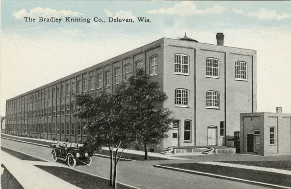 Colored postcard of an illustration of a three-story industrial building on the other side of the street. There is an automobile driving by. Caption reads: "The Bradley Knitting Company, Delavan, Wis."