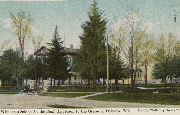 Hand-colored view of the drive and front gate of the Wisconsin School for the Deaf. The main building is obscured by trees. Caption reads: "Wisconsin School for the Deaf, Approach to the Grounds, Delavan, Wis."