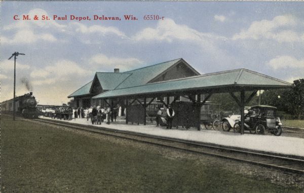 Hand-colored view across railroad tracks towards a train depot, with passengers gathered on the platform awaiting an arriving train. Horse-drawn wagons and automobiles are parked behind the platform. Caption reads: "C. M. and St. Paul Depot, Delavan, Wis."