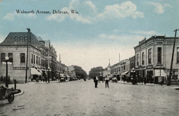 Hand-colored view down center of a commercial street lined with buildings and street lamps. Two men are standing in the center of the intersection. Automobiles are parked along the curbs. A hardware store is on the left corner; and a theater is on the right corner. Caption reads: "Walworth Avenue, Delavan, Wis."