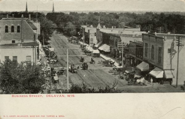 Elevated view of a commercial street. Horses, wagons and buggies are parked at the curbs and moving down the street. Church steeples are in the far background beyond trees. Caption reads: "Business Street, Delavan, Wis."