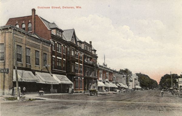 View down center of unpaved street towards commercial buildings on the left. There are pedestrians, and horse-drawn vehicles on the sidewalks and along the curb. Caption reads: "Business Street, Delavan, Wis."