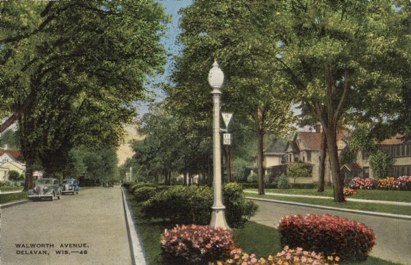 View down wide median in a tree-lined residential neighborhood. The median is landscaped with flowers and bushes, and a lamppost. Automobiles are parked on the left along the curb. Caption reads: "Walworth Avenue, Delavan, Wis."