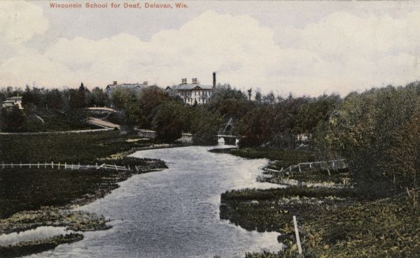 Elevated view of a river with grassy and tree-lined banks. The Wisconsin School for the Deaf is on a hill in the far distance beyond a bridge over the river. Caption reads: "Wisconsin School for the Deaf, Delavan, Wis."