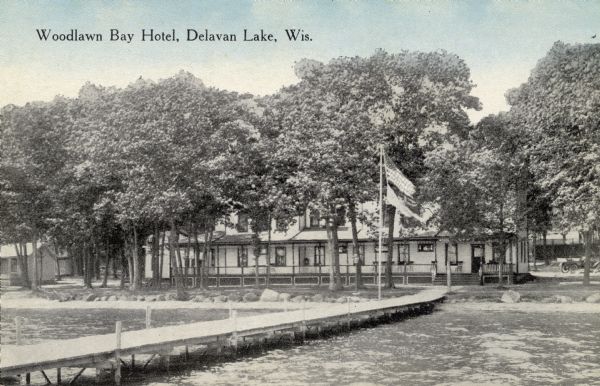 View across Delavan Lake towards the Woodlawn Bay Hotel surrounded by trees. There is a long pier extending from the shoreline towards the left. At the shoreline is a flag pole with two flags. Caption reads: "Woodlawn Bay Hotel, Delavan Lake, Wis."