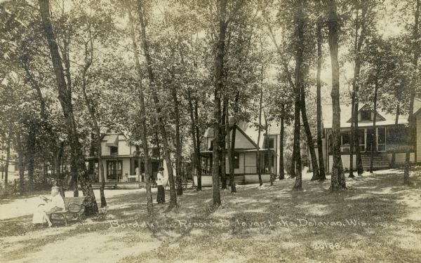 View of the tree-filled grounds, and a few cabins, at the Bord du Lac Resort. Two people are sitting on a bench in the foreground on the left. Caption reads: "Bord du Lac Resort, Delavan Lake, Delavan, Wis."