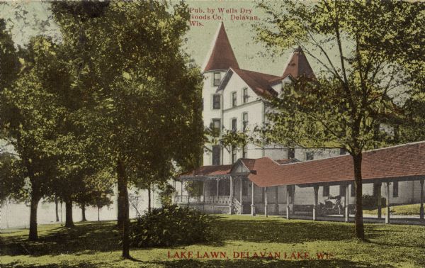 Hand-colored view of the Lake Lawn Resort. Delavan Lake is in the background on the left. There is an automobile parked under a roofed entryway that leads to the front porch. Caption reads: "Lake Lawn, Delavan Lake, Wis."