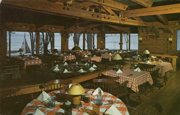Photographic postcard view of the dining room at Lake Lawn Lodge. The ceiling has exposed beams, and the tables are set with tableware. There is a view of the lake from the large windows in the background.
