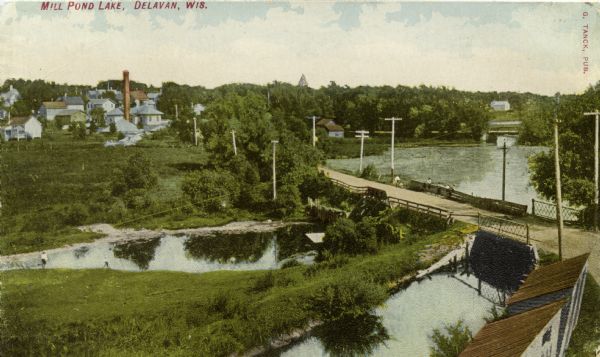 Elevated view of Mill Pond Lake. There is a road and bridge crossing an outlet in the foreground. Houses and other buildings are across the lake in the background. Caption reads: "Mill Pond Lake, Delavan, Wis."
