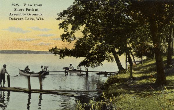 Hand-colored photographic postcard view along the shoreline at Delavan Lake. There is a person standing on one of the two piers, and four more people are sitting in two rowboats. Caption reads: "View from the Shore Path at Assembly Grounds, Delavan Lake, Wis."