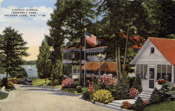Hand-colored photographic postcard of a house-lined street leading down to a lake. Flowers, bushes and trees are planted along the curb. Caption reads: "Lincoln Avenue, Assembly Park, Delavan Lake, Wis."