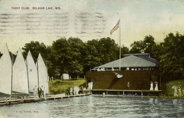 View across lake towards a line of sailboats at the dock of the Yacht Club. People are standing on the pier, and on the boathouse.