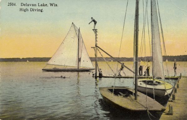 View from shoreline of a high diver standing on a platform at the end of a pier on Delavan Lake. People are standing on the pier watching the diver, and a few people are swimming in the water. Sailboats are docked at the pier, and one is anchored just beyond the pier. Caption reads: "Delavan Lake, Wis. High Diving."