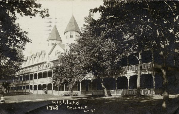 Black and white photographic postcard view across lawn towards a large hotel with a long porch, long balcony and two spires. Caption reads: "Highland, Delavan Lake."