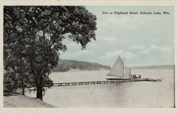 View from shoreline towards the Highland Hotel Pier. A group of people are standing at the end of the long pier near a sailboat docked at the side. A high dive platform is on the end of the pier. Caption reads: "Pier at Highland Hotel, Delavan Lake, Wis."