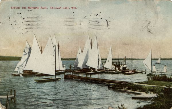 View from shoreline towards several small sailboats, and a boat moored around two piers. People are standing on the piers. Caption reads: "Before the Morning Race, Delavan Lake, Wis."