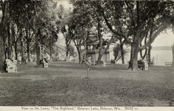 View of the Highland Hotel's lakeside lawn. Several park benches are among the trees on the lawn. In the background at the shoreline is a boathouse with a roofed porch on top, and a long, wooden water slide.