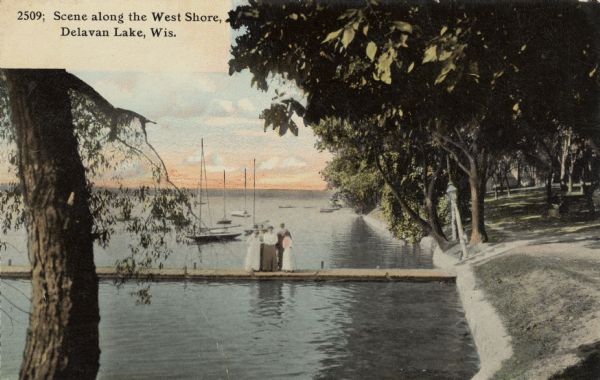 View along shoreline towards a group of people standing on a pier. Sailboats are anchored in the water in the background. Caption reads: "Scene Along the West Shore, Delavan Lake, Wis."