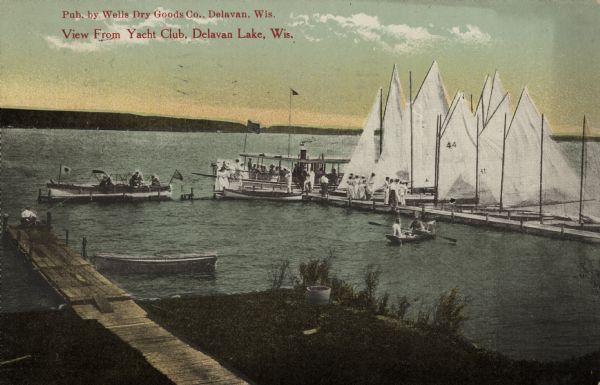 Elevated view looking down towards two piers in Delavan Lake. A group of people, several sailboats and an excursion boat are at the pier on the right. There is a rowboat and a person fishing on the pier on the left. Caption reads: "View From Yacht Club, Delavan Lake, Wis."