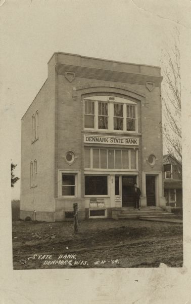 Photographic postcard view of the bank building, with a man standing on the steps at the entrance. Caption reads: "State Bank, Denmark, Wis."