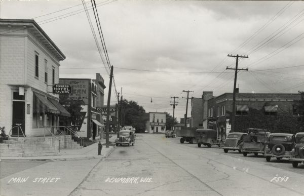Photographic postcard view of the Main Street commercial district, with automobiles and trucks parked at the curbs. Signs are on the buildings for Dinner's Hotel Tavern, Collard's Bar and Denmark Bakery. Caption reads: "Main Street, Denmark, Wis."
