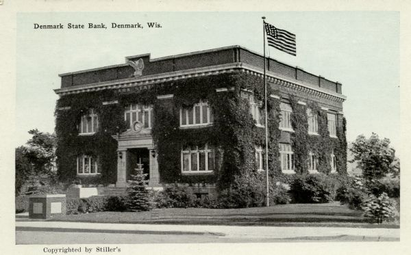 View from street towards the Denmark State Bank, which is heavily covered with ivy. There is a flagpole at the corner flying the American flag. Caption reads: "Denmark State Bank, Denmark, Wis."