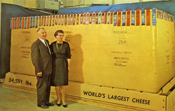 Colored postcard view of Mr. & Mrs. Steve Siudzinski, owners of Steve's Cheese, in front of a 34,591 pound block of cheddar cheese draped with prize winning ribbons that the company made. This famous cheese was on display at the New York World's Fair.