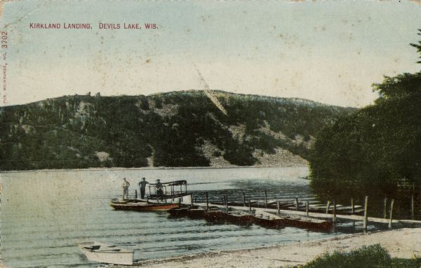 View from shoreline of boats, including an excursion boat, on the shoreline and along both sides of a pier. There are three men on the pier at Kirkland Landing at Devil's Lake. Caption reads: "Kirkland Landing, Devils [sic] Lake, Wis."