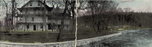 Color enhanced black and white photographic postcard, a double length panorama of the Lake View Hotel. The lake shore is stabilized with a rock wall and a motorboat is on the shoreline.