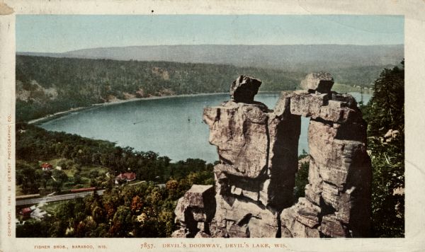 Colorized postcard of an elevated view of the Devil's Doorway rock formation at Devil's Lake State Park. Devil's Lake is below in the background. Caption reads: "Devil's Doorway, Devil's Lake, Wis."