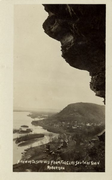 Black and white photographic postcard view of De Soto, taken from the cliff south of town, looking north along the Mississippi River. Caption reads: "A View of DeSoto, Wis. from the Cliff South of Town."