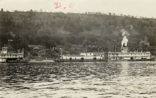 Black and white photographic postcard view across water towards three steamboats on the shoreline of the Mississippi River.