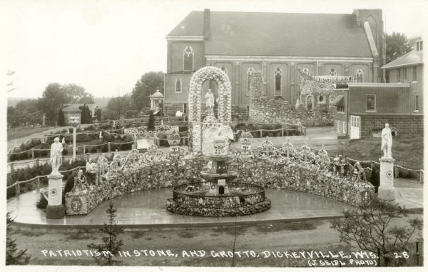 Photographic postcard view of the grounds of the Dickeyville Grotto. The sculpture, which was created by Father Matthias Wernerus in 1929-30, and honors Columbus, Washington and Lincoln. In the center of the scene is a fountain with the sculpture of an eagle on the top. In the background is the Holy Ghost Church. Caption reads: "Patriotism in Stone, and Grotto, Dickeyville, Wis."