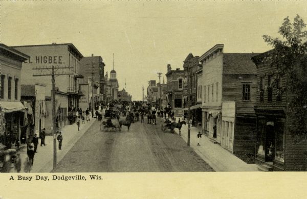 Elevated view of a busy street scene on Iowa Street, looking north. The Higbee Hotel is on the left side of the street, and the Iowa County Courthouse is in the background. Caption reads: "A Busy Day, Dodgeville, Wis."