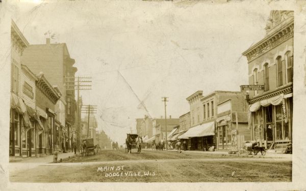 View down center of unpaved Main Street, looking north, showing business storefronts and horse-drawn buggies. The Iowa County Courthouse is in the background. Caption reads: "Main St., Dodgeville, Wis."