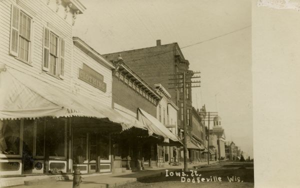 Photographic postcard view of Iowa Street looking north, showing businesses, with awnings over the show windows, on the west side of street. The Iowa County Courthouse is in the background. Caption reads: "Iowa St., Dodgeville, Wis."
