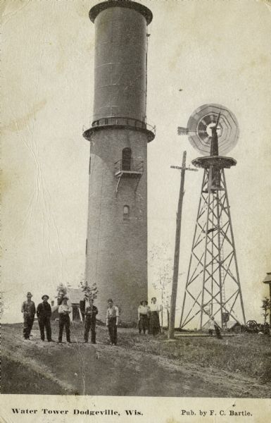 View of a group of people posing in front of the water tower. There is a windmill nearby on the right side. Caption reads: "Water Tower, Dodgeville, Wis."