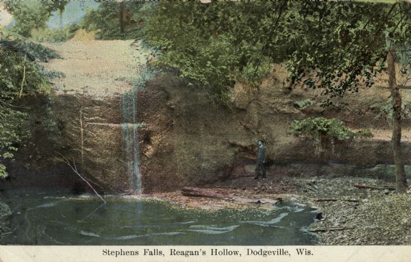 Color enhanced postcard across water towards a man standing near a cliff face and observing a waterfall. Located in Governor Dodge State Park on STH 23 north of Dodgeville. Caption reads: "Stephens Falls, Reagan's Hollow, Dodgeville, Wis."