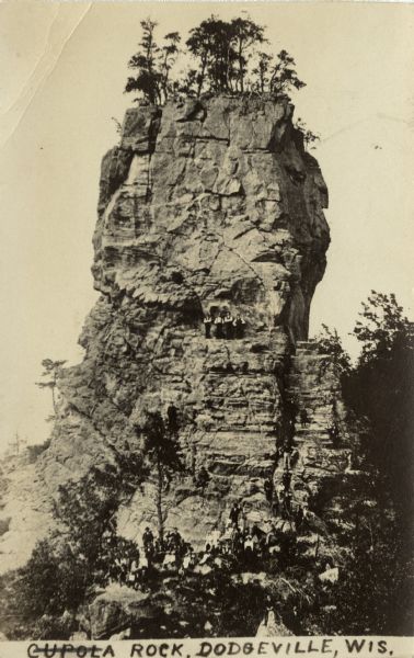 Elevated view of people posing at the base, and also part of the way up, Cupola Rock. Now called Percussion Rock. The rock formation is located north of Dodgeville, west of STH 23, on Percussion Rock Road. Caption reads: "Cupola Rock, Dodgeville, Wis."