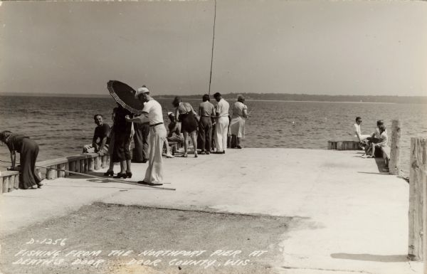 Photographic postcard view looking out at people standing, and some fishing, on the Northport pier at Death's Door, with the bay in the background.