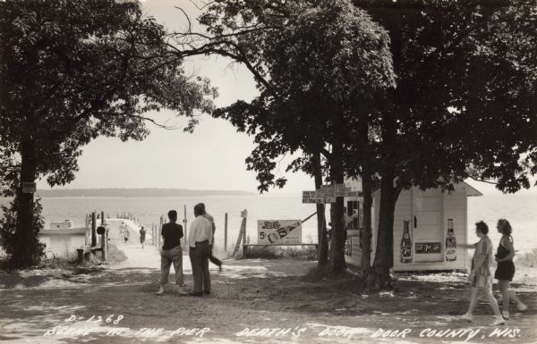 Black and white photographic postcard of people on the shoreline and on the pier at Death's Door. Signs on a tree near a snack stand on the right read: "Smoked Fish", "Speed Boat Rides", "Gills Rock Ferry Dock", and "Night Crawlers for Sale". Caption reads: "Scene at the Pier Death's Door Door County, Wis."
