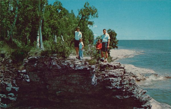 View across water towards a man, woman, and two children standing on the rock ledge at Cave Point. Lake Michigan is on the right.