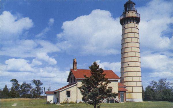 Color photographic postcard view of the Cana Island U.S. Lighthouse surrounded by a green lawn. There is a hand pump near the building.