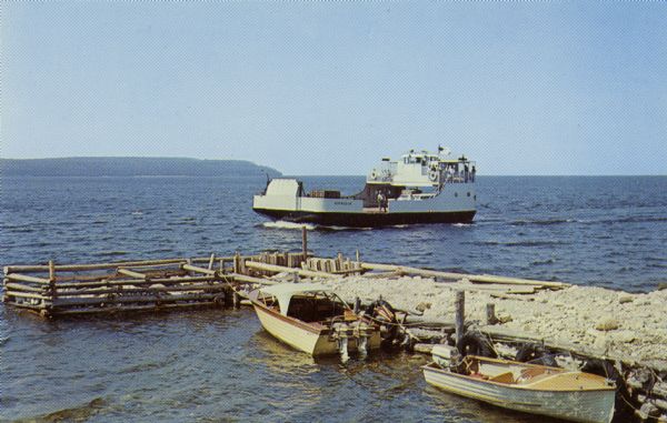 Color postcard view of the "Voyageur," a car and passenger ferry operating between mainland Door County and Washington Island. Two motorboats are moored on a pier at the shoreline.