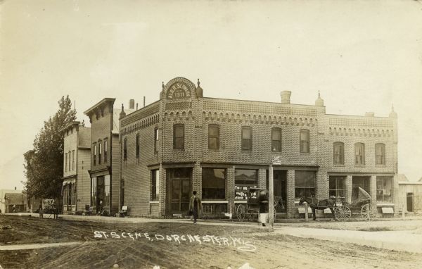 View from unpaved street towards the Alb. R. Schwahn building on a corner. There are pedestrians on the sidewalk, and horse-drawn vehicles are parked near the curbs. Caption reads: "St. Scene, Dorchester, Wis."