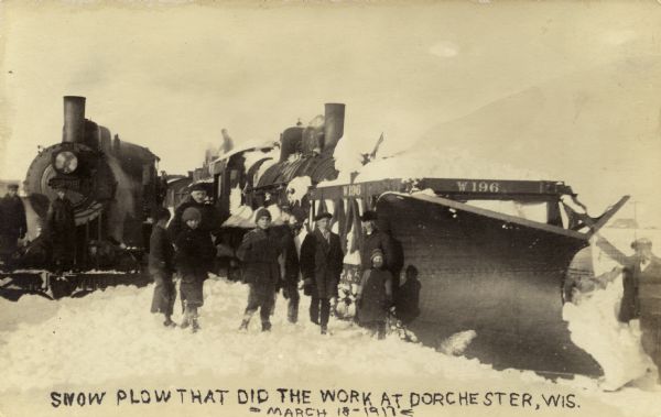 Photographic postcard view of a group of people standing in front of two locomotives, the one on the right with a snowplow attached. Caption reads: "Snow Plow That Did the Work at Dorchester, Wis."