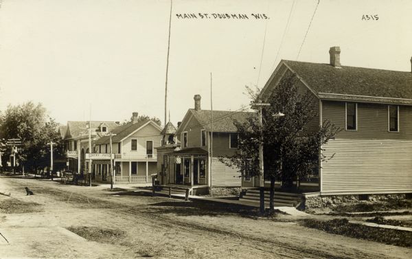 Slightly elevated view looking across the unpaved main street towards wooden buildings along a sidewalk. Caption reads: "Main Street, Dousman, Wis."